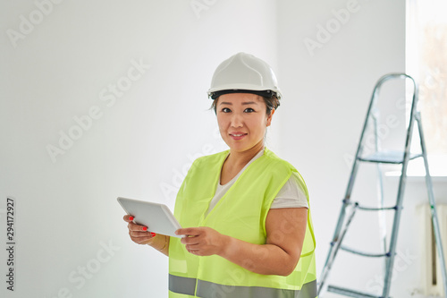 Repairs in the apartment, a girl in a green vest and a white helmet is in a white room with a tablet in her hands, she makes repairs in the background is a ladder