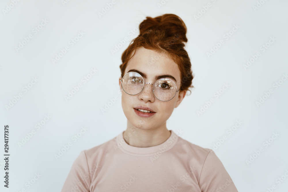 Charming young girl with curly red hair braided in the bun on her head, with freckles on her face with glasses on a white background in the pale pink T-shirt cute smiles Charming young girl with curly red hair braided in the bun on her head, with freckles on her face with glasses on a white background in the pale pink T-shirt cute smiles