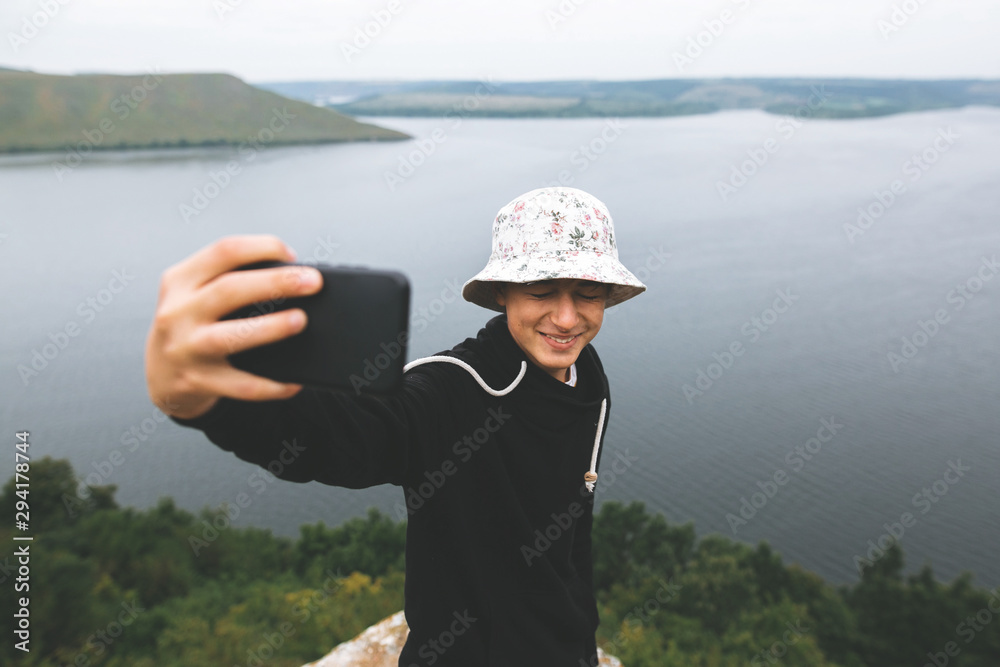 Hipster teenager taking selfie on phone and smiling, standing on top of rock mountain with amazing view on river. Young stylish guy exploring and traveling. Atmospheric moment. Copy space