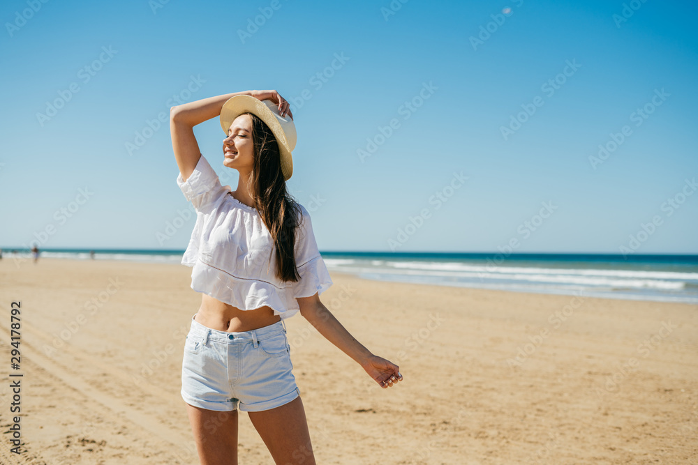 Gorgeous young girl with long dark hair and the straw hat on her head in a white sweater and shorts stands on the background of the sea coast and is surprised Gorgeous young girl with long dark hair and the straw hat on her head in a white sweater and shorts stands on the background of the sea coast and is surprised