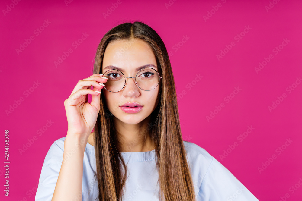 Strict young girl with long dark hair and glasses on a pink background is standing and looking up