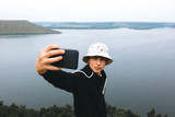 Hipster teenager taking selfie on phone while standing on top of rock mountain with amazing view on river. Young stylish guy exploring and traveling. Atmospheric moment. Copy space