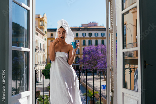 Young girl with a white towel on her head stands on the balcony, eats and drinks coffee from a blue mug