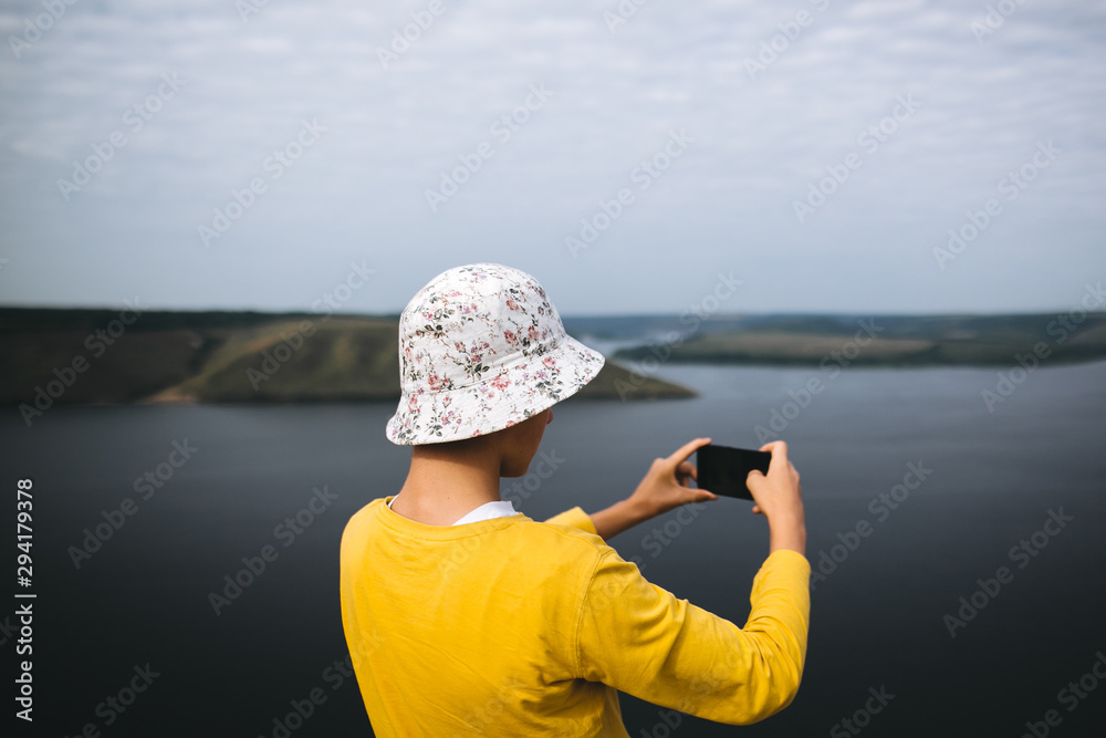 Hipster teenager streaming live while standing on top of rock mountain with amazing view on river. Young stylish guy exploring and making video of travel. Copy space