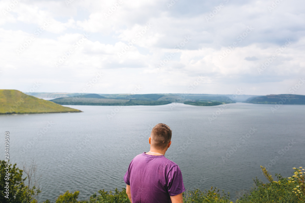 Stylish hipster traveler in purple shirt standing on top of rock mountain with amazing view on river. Young brutal guy exploring and traveling, back view. Copy space