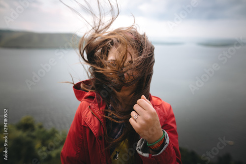 Traveler girl in red raincoat and with windy hair standing on top of rock mountain, enjoying beautiful view on river. Young hipster woman in glasses relaxing on cliff. Copy space. Explore photo