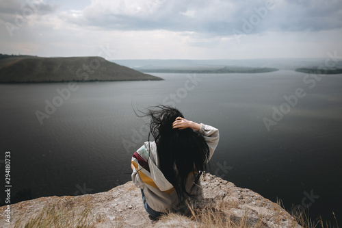 Back of stylish hipster girl sitting on top of rock mountain with beautiful view on river. Young tourist woman with windy hair relaxing on cliff. Atmospheric moment. Copy space