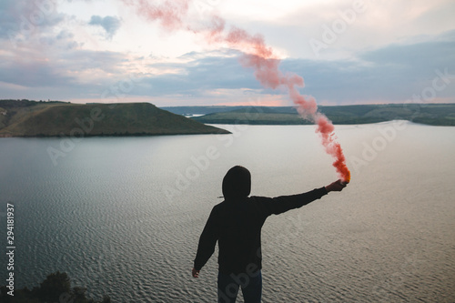 Stylish hipster holding red smoke bomb in hand, standing on top of rock mountain with amazing sunset view on river. Atmospheric moment. Traveler guy with smoke. Copy space