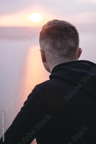 Hipster traveler enjoying amazing sunset view on river while standing on top of rock mountain. Brutal guy in windbreaker exploring and traveling. Atmospheric moment. Copy space