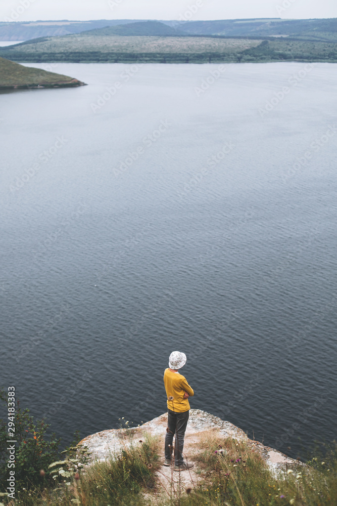 Hipster teenager sitting  on top of rock mountain and looking on river, view from back. Young stylish guy exploring and traveling. Atmospheric tranquil moment. Copy space