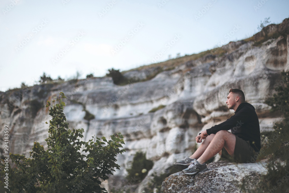 Hipster traveler sitting on top of rock mountain and enjoying amazing view on river. Stylish guy in windbreaker exploring and traveling. Atmospheric tranquil moment. Copy space