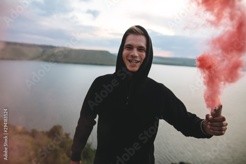 Stylish hipster holding red smoke bomb in hand, standing on top of rock mountain with amazing sunset view on river. Atmospheric moment. Traveler guy with smoke. Copy space