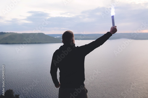 Obraz na płótnie Brutal man in black windbreaker holding blue flare torch in hand, standing on top of rock in evening with view on river
