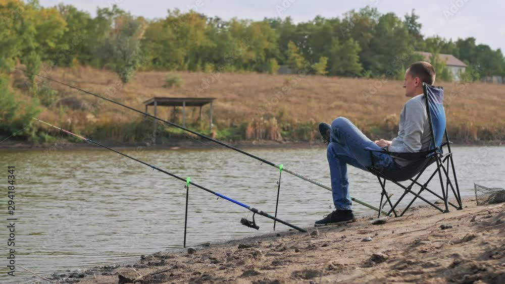 A man is fishing in the lake. He is sitting on a chair, waiting for the fish to peck. Camping and fishing. Real time 4k footage. A man is fishing in the lake. He is sitting on a chair, waiting for the fish to peck. Camping and fishing. Real time 4k footage.