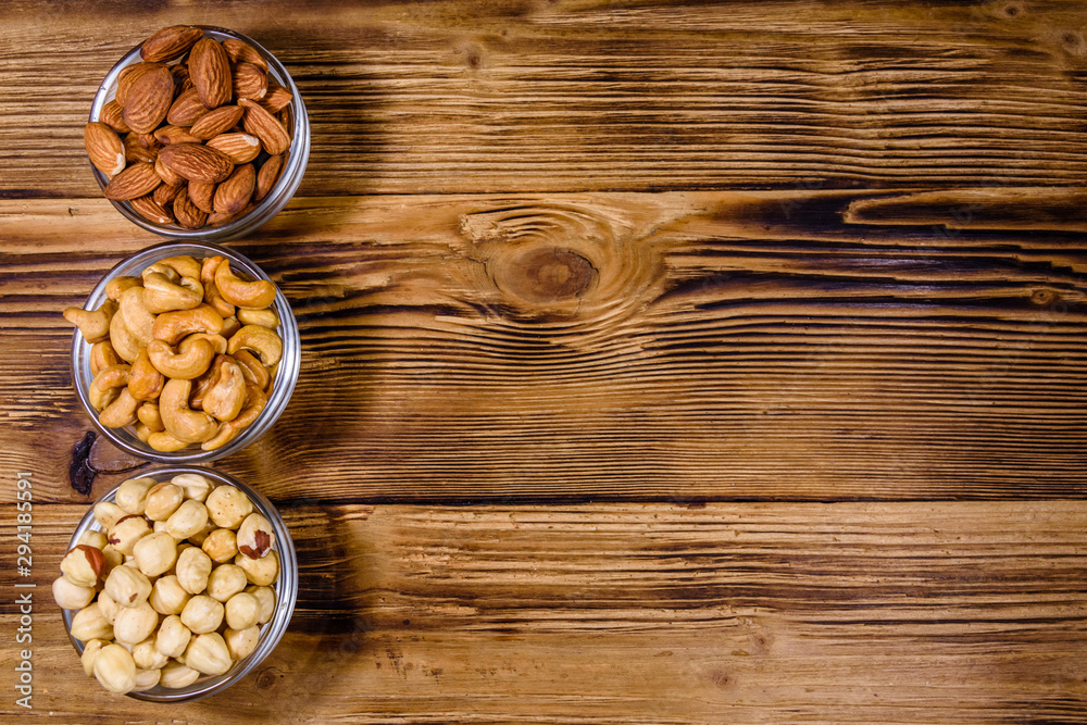 Naklejka premium Various nuts (almond, cashew, hazelnut) in glass bowls on a wooden table. Vegetarian meal. Healthy eating concept. Top view