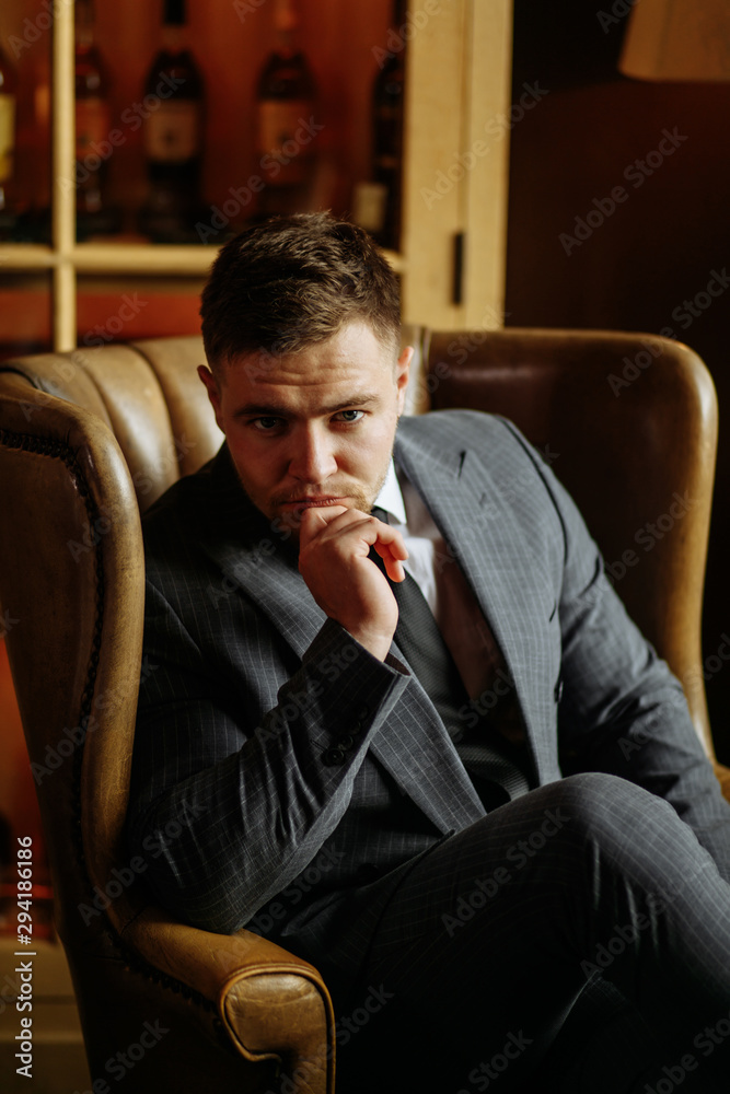 Young brave man with crossed legs sitting in beautiful armchair, keeping chin, looking cautiously towards camera, against background of well designed study Young brave man with crossed legs sitting in beautiful armchair, keeping chin, looking cautiously towards camera, against background of well designed study