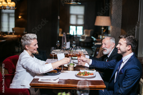 Happy business partners cheerfully raising hands with glasses of wine, gladly congratulate each other with successful deal, posing in luxury restaurant