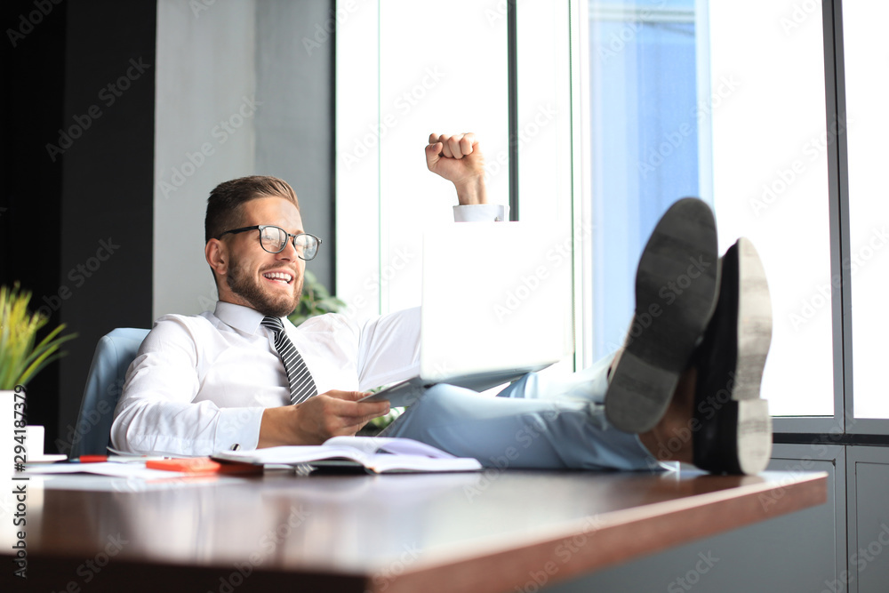 Handsome businessman sitting with legs on table and keeping arm raised and expressing joyful in office Handsome businessman sitting with legs on table and keeping arm raised and expressing joyful in office
