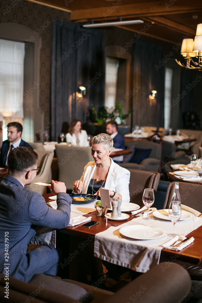 Charming blonde well dressed woman with short haircut actively talks to male friend, shows cheerful expression at setted table in luxury restaurant