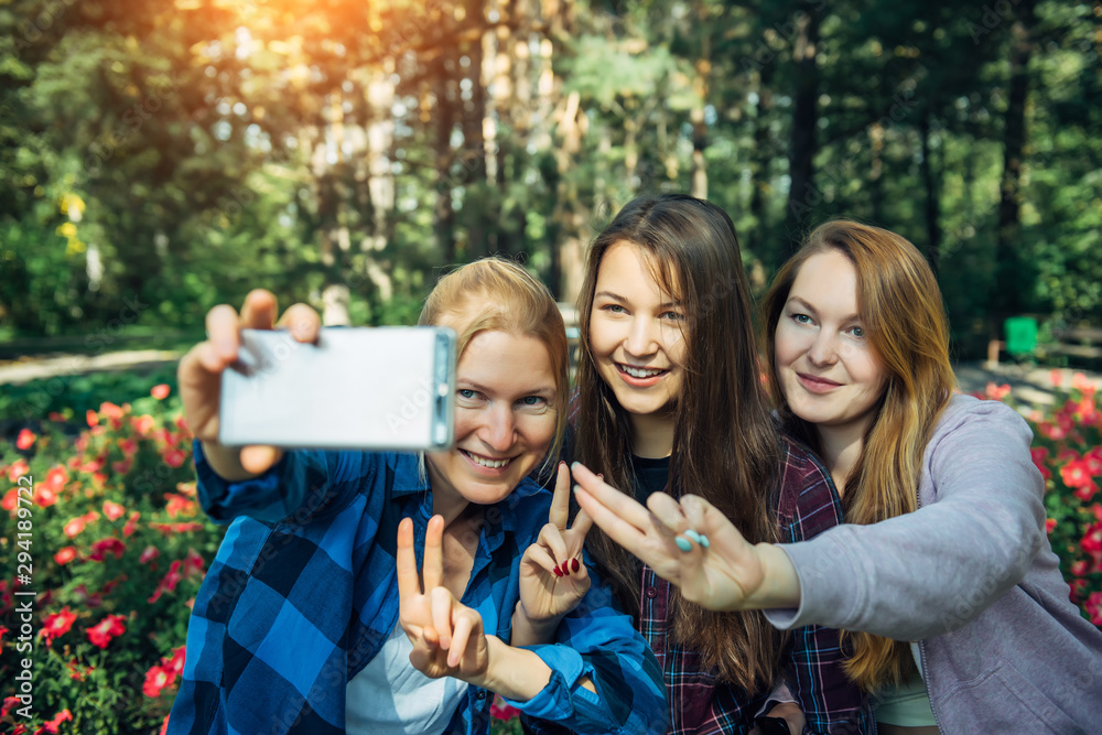 Fototapeta premium Portrait of cute girlfriends taking selfie with smartphone in summer park. Friendship, technology and people concept - happy young women having fun and taking photos for instagram.
