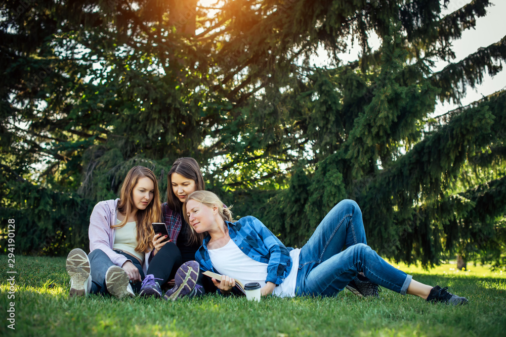 Three young girls sitting on green grass with book and smartphones. Cheerful friends resting in park. Spring and summer leisure on a nature, concept of female friendship. Three young girls sitting on green grass with book and smartphones. Cheerful friends resting in park. Spring and summer leisure on a nature, concept of female friendship.