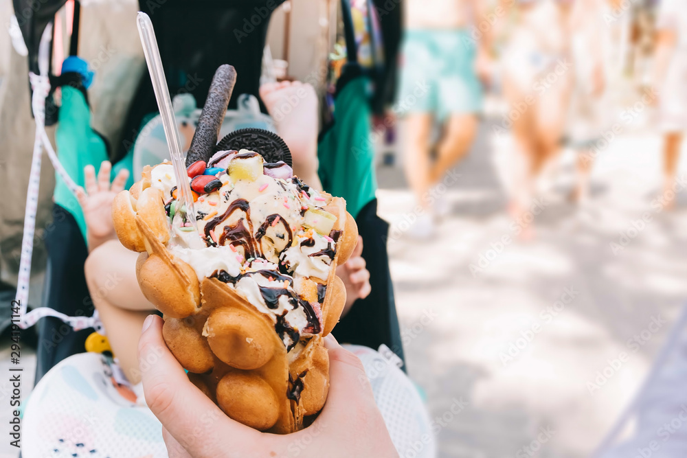 A girl holds a Hong Kong waffle dessert in her hand. Sweets with ice cream, cookies, syrup and sweets. Against the background of a child in a stroller.