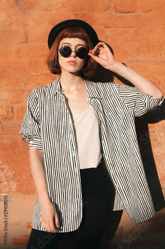Young woman in striped shirt, hat and sunglasses posing near the orange wall