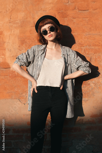 Young woman in striped shirt, hat and sunglasses posing near the orange wall