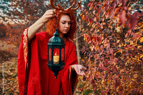 Woman in long red dress with deer horns in autumn forest. A female in the fall forest walks with a lantern and looking for magic herbs. The concept of a fairy tale.