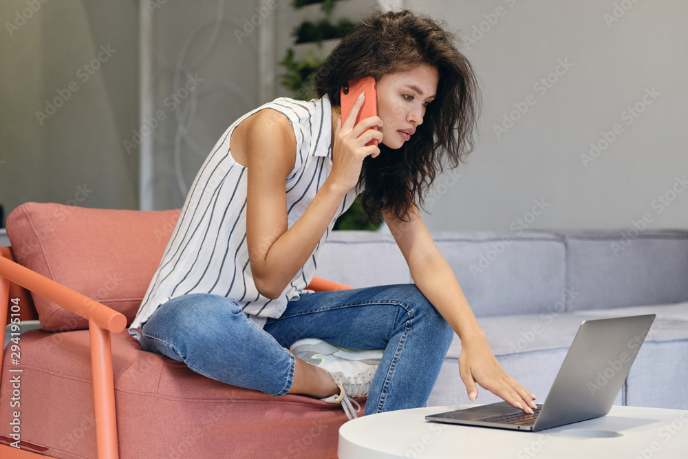 Young beautiful casual asian woman talking on cellphone and thoughtfully working on laptop in co-working space Young beautiful casual asian woman talking on cellphone and thoughtfully working on laptop in co-working space