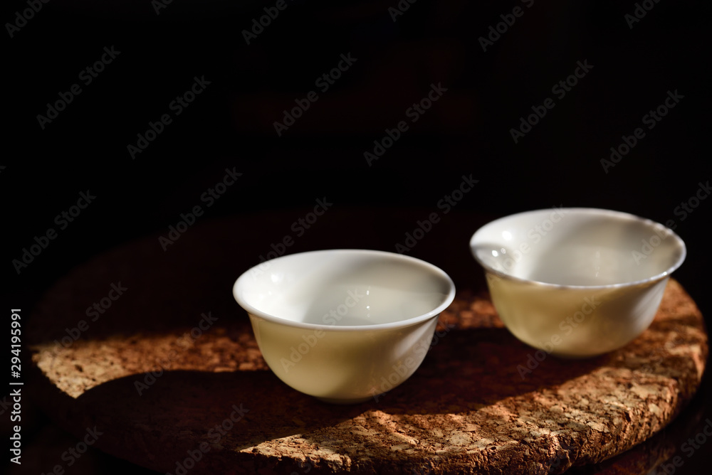 Close up of two empty small white chinese tea cups standing against a dark background on a cork plate with free space for text at a Chinese tea ceremony Close up of two empty small white chinese tea cups standing against a dark background on a cork plate with free space for text at a Chinese tea ceremony