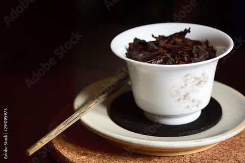 Closeup of a typical Chinese porcelain teapot in white with brewed black tea leaves in it on a saucer and a cork plate with an old wooden stick against a dark background at a tea ceremony