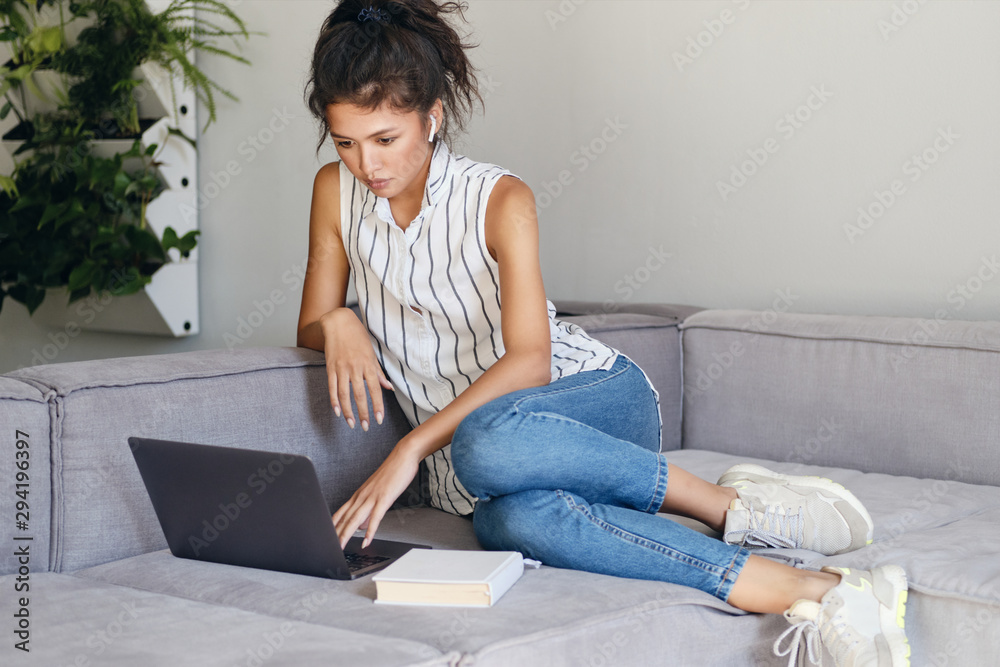 Beautiful confident Asian girl thoughtfully working on laptop on sofa at home