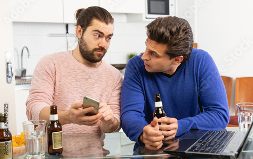 Two cheerful men talking and laughing while enjoying beer at home, using phone