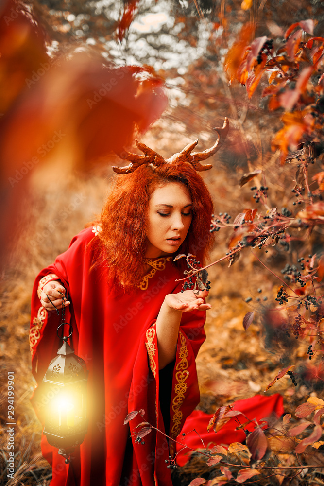 Naklejka premium Woman in long red dress with deer horns in autumn forest. A woman in the autumn forest walks with a lantern and looking for magic herbs. The concept of a fairy tale.