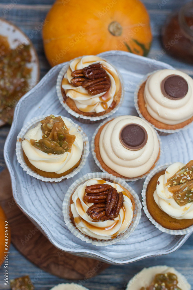Pumpkin cupcakes with cream cheese frosting decorated with pecan nuts, maple syrup, caramel bites and chocolate toffee candies on wooden rustic background Pumpkin cupcakes with cream cheese frosting decorated with pecan nuts, maple syrup, caramel bites and chocolate toffee candies on wooden rustic background