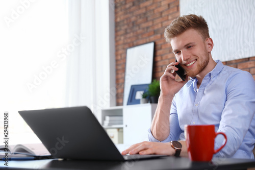 Young man talking on phone while using laptop at table indoors