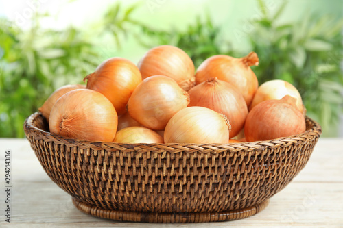 Wicker basket with ripe onions on white wooden table against blurred background