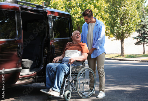 Foto Young man helping patient in wheelchair to get into van outdoors