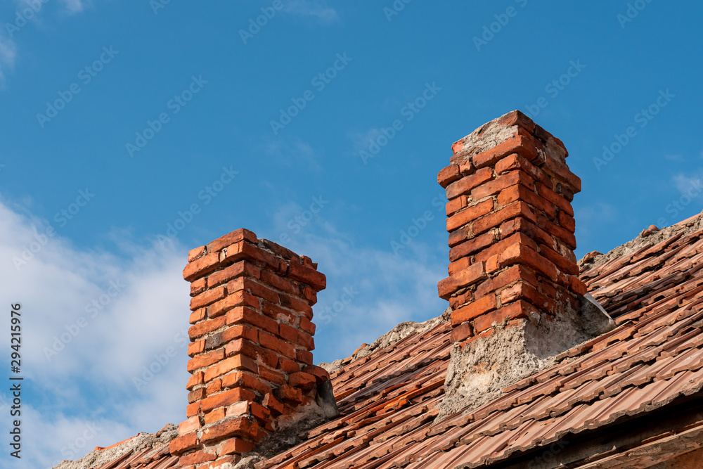 Two traditional red brick chimneys on an old clay tile roof against a blue sky. Chimney in need of sweeping and repair. Preparing for winter concept.