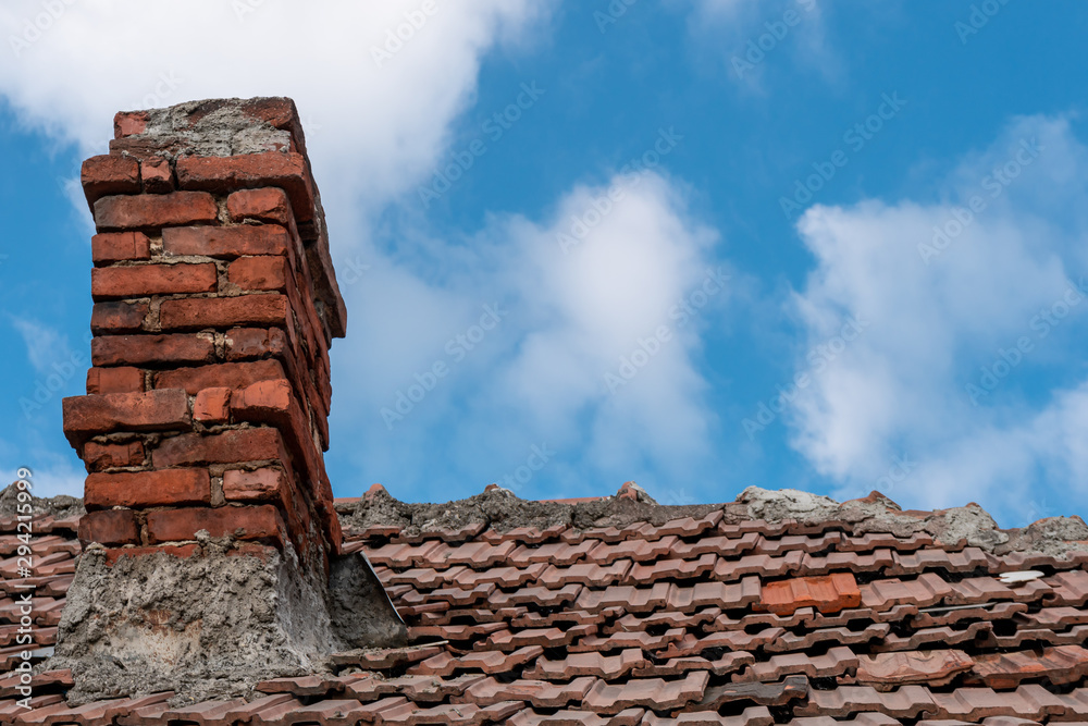 Naklejka premium Old red terracotta tile roof with a damaged traditional large brick chimney in need of repair, on a blue sky background. Home winter preparations.