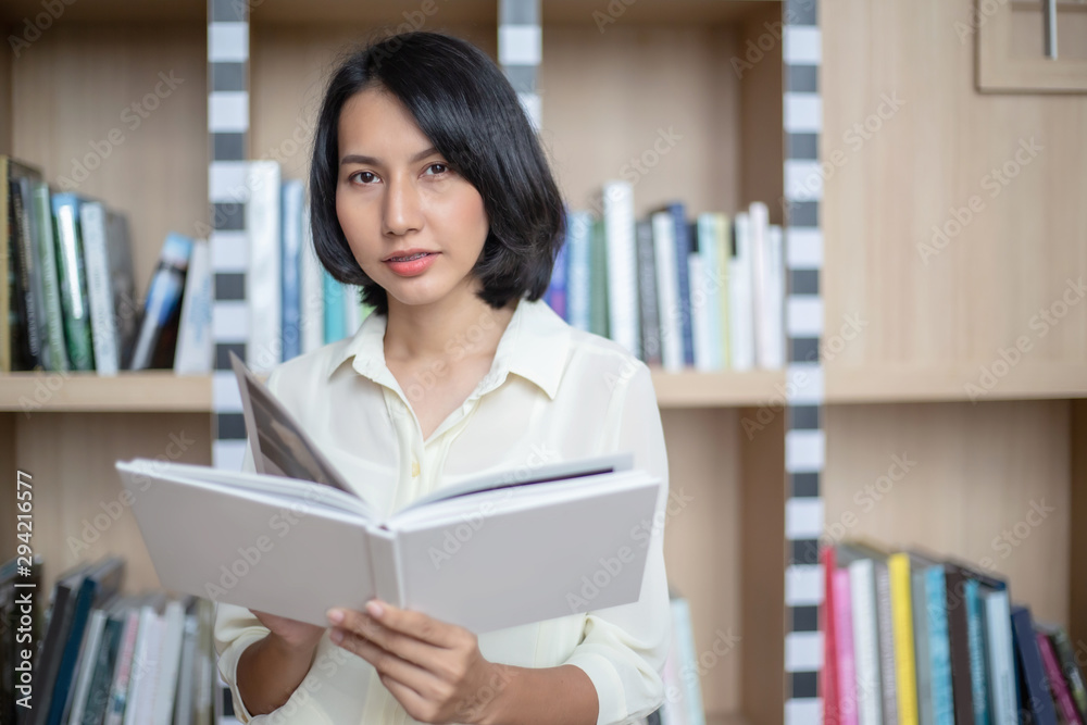 Young and beautiful businesswoman holding a book in her hands and reading at the bookshelves in the modern office Young and beautiful businesswoman holding a book in her hands and reading at the bookshelves in the modern office