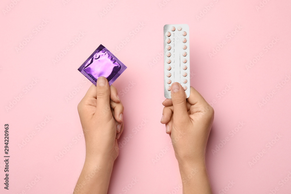 Woman holding condom and birth control pills on pink background, top view. Safe sex - obrazy, fototapety, plakaty 