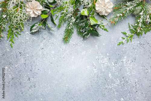 Christmas or winter background with a border of green and frosted evergreen branches and pine cones on a grey vintage board. Flat lay