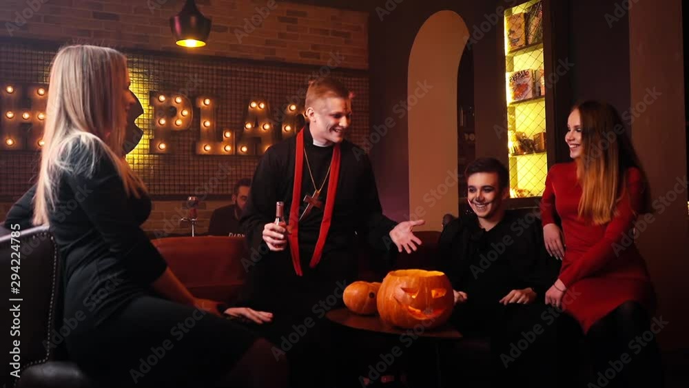Bunch of friends have fun and carefree time celebrating Halloween. Group of young people sitting in cafe in festive costumes and celebrating an autumn holiday. Holiday carved pumpkin on the table.