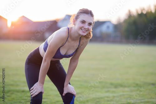 Slim blonde girl plays sports and performs yoga poses in summer grass covered stadium on a sunset background. Woman doing exercises on the yoga mat. © bilanol