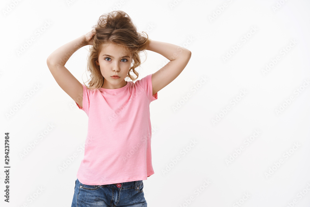 Glamour young cute blond little girl child with blue eyes in pink t-shirt raising hair up, making hair-bun, look tender, fashion gaze camera, serious determined expression, stand white background Glamour young cute blond little girl child with blue eyes in pink t-shirt raising hair up, making hair-bun, look tender, fashion gaze camera, serious determined expression, stand white background