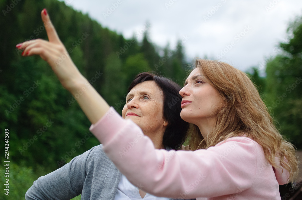 Happy senior mother and adult daughter are travelling and posing together over landscape of forest and mountains, young girl is pointing at something. Concept of travelling with family Happy senior mother and adult daughter are travelling and posing together over landscape of forest and mountains, young girl is pointing at something. Concept of travelling with family