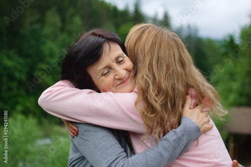 Outdoors portrait of happy smiling mother and daughter who are embracing each other outside over landscape of forest and mountains.