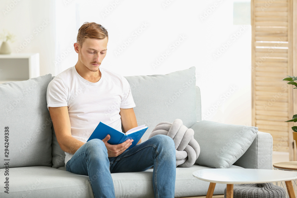 Young man in stylish t-shirt and with book sitting on sofa at home